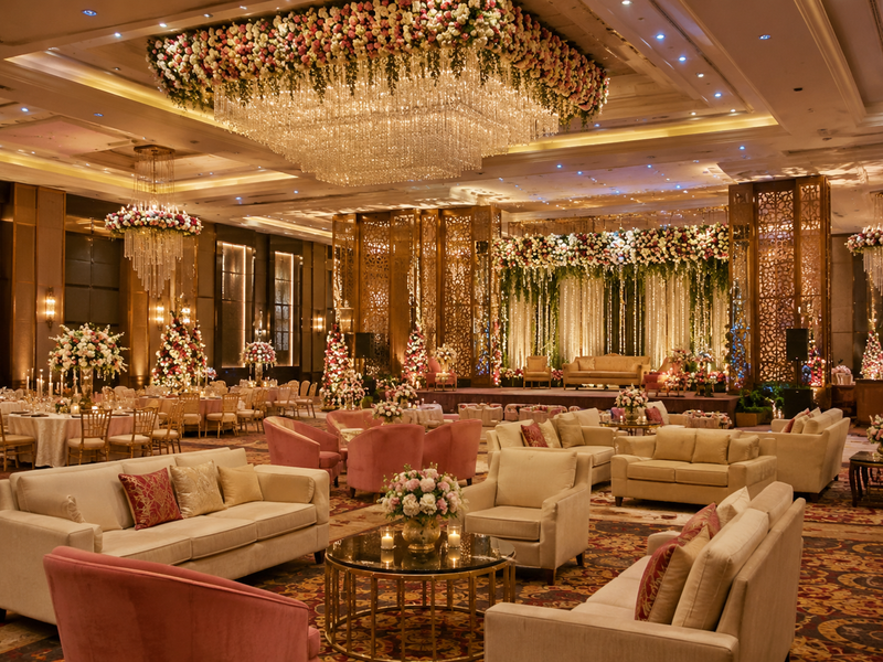 Elegant banquet hall decoration featuring luxury sofas, floral stage backdrop, stylish seating arrangements, chandeliers, and warm lighting for engagement and reception parties.